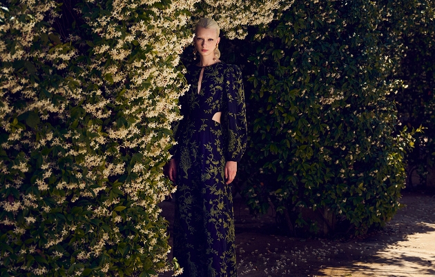 Gmodel in green floral and black dress standing inbetween hedges